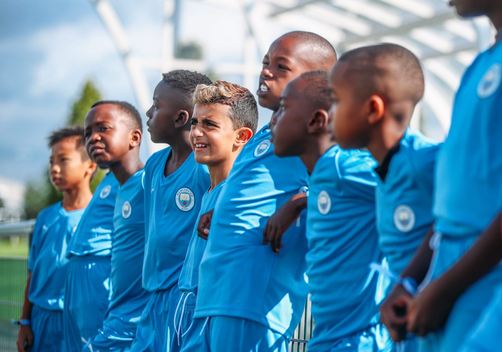A group of young boys wearing a Manchester City Football kit, leaning against a railing. One boy is looking ahead and smiling.