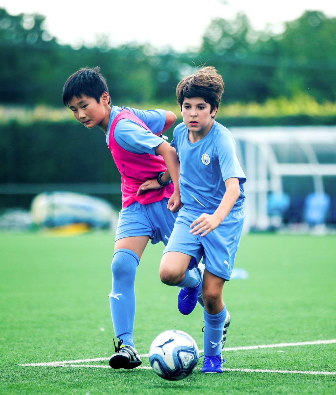 Two boys running after a football. They are both wearing a Manchester City Football School kit. One boy is pushing the other boy out of the way in order to reach the ball.