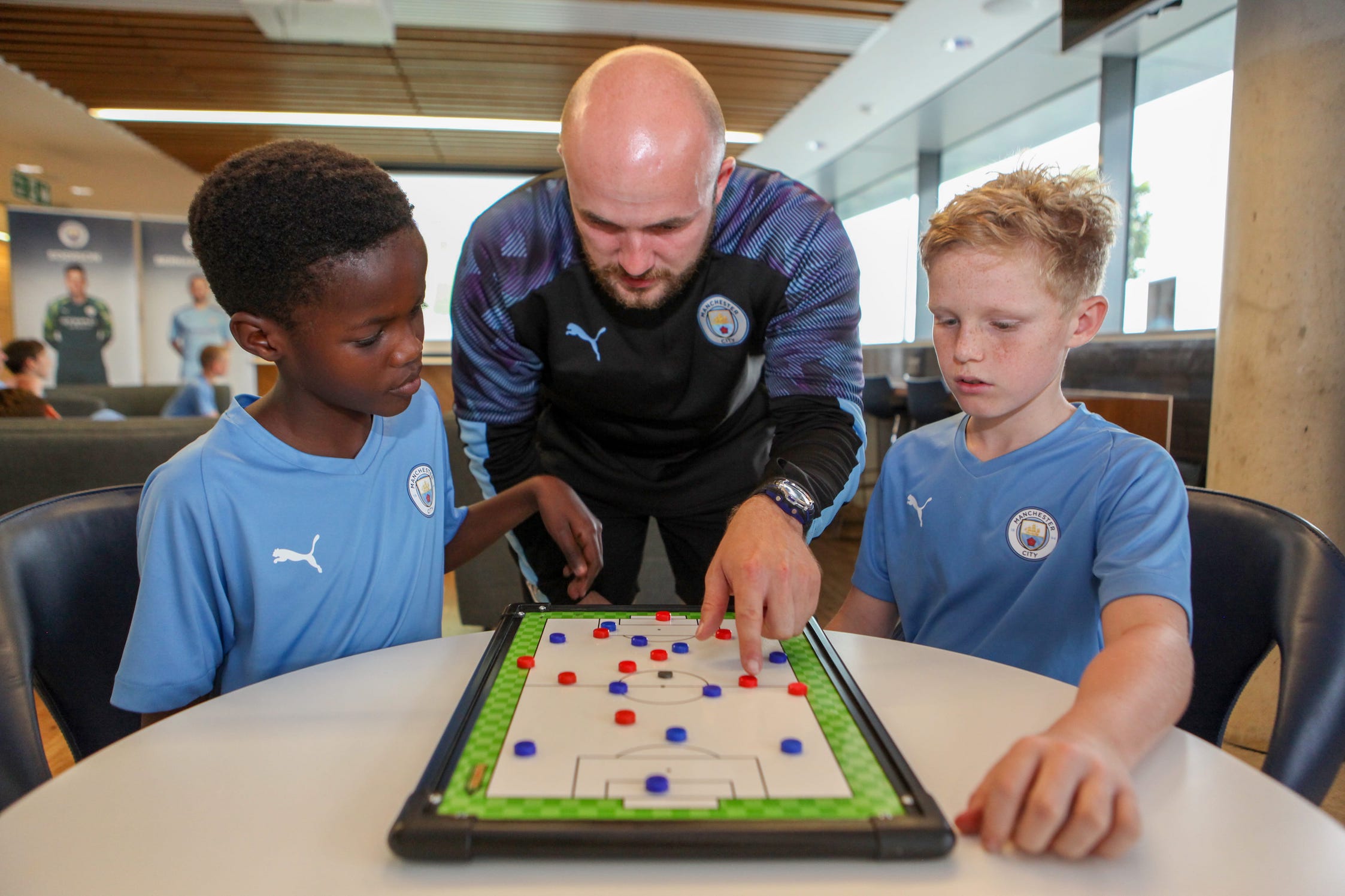 Two young boys on the Football Development Programme at Manchester City Football School. They are sat looking at a whiteboard on the table in front of them whilst their coach is crouching down in between them and pointing at the board.