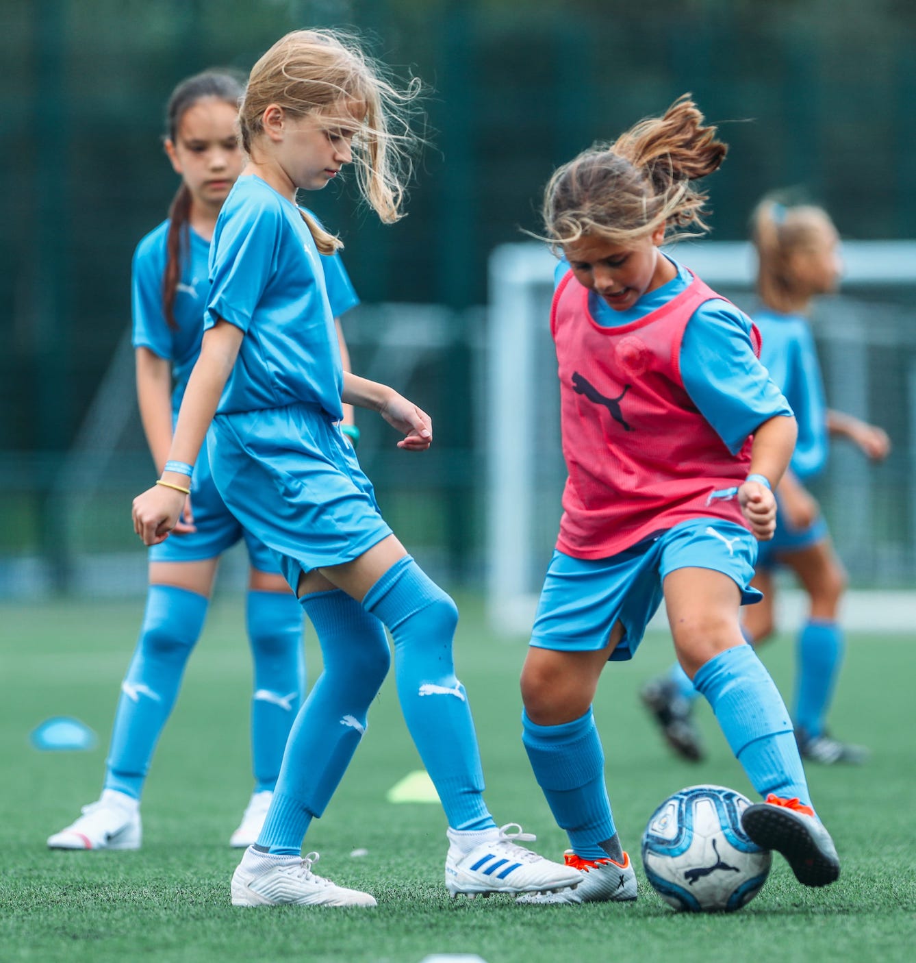 Two young girls playing football at the City Football Academy. One girl is wearing a red bib and is in possession of the ball.