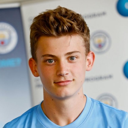 A young boy with dark hair and braces is posing for a photo. He is wearing a Manchester City Football School top. The background is blurred.