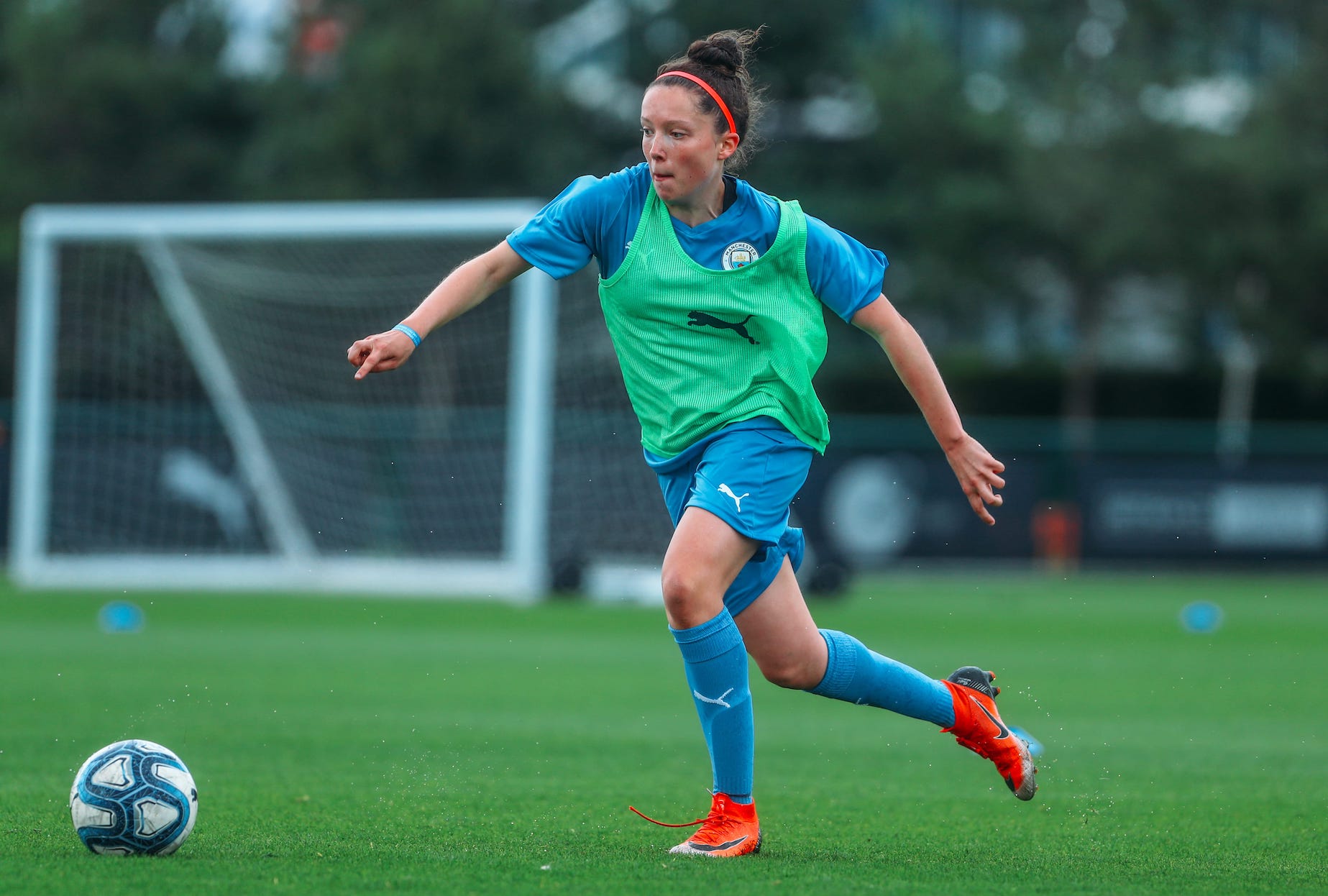 A female footballer running towards a ball. She is wearing a blue, Man City Football School kit and a green bib.