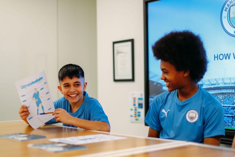 Two young boys on the Language Course at City Football Language School. They are both sitting at a table and wearing a blue football kit. One boy is holding a piece of paper and smiling while the other boy looks at him.