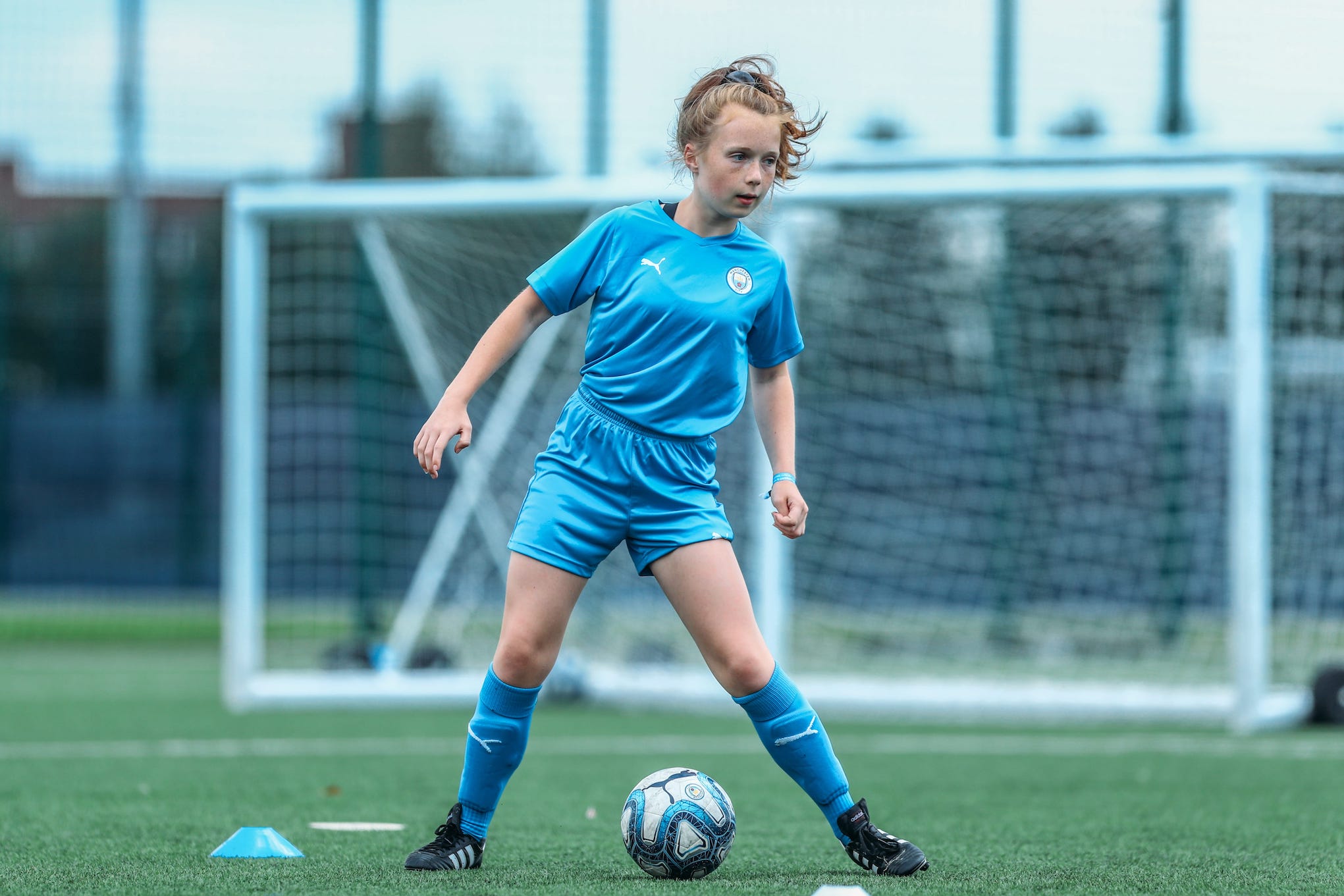 A young girl with blond hair on a football pitch. She is wearing a Manchester City Football School kit. There is a goal post in the background.