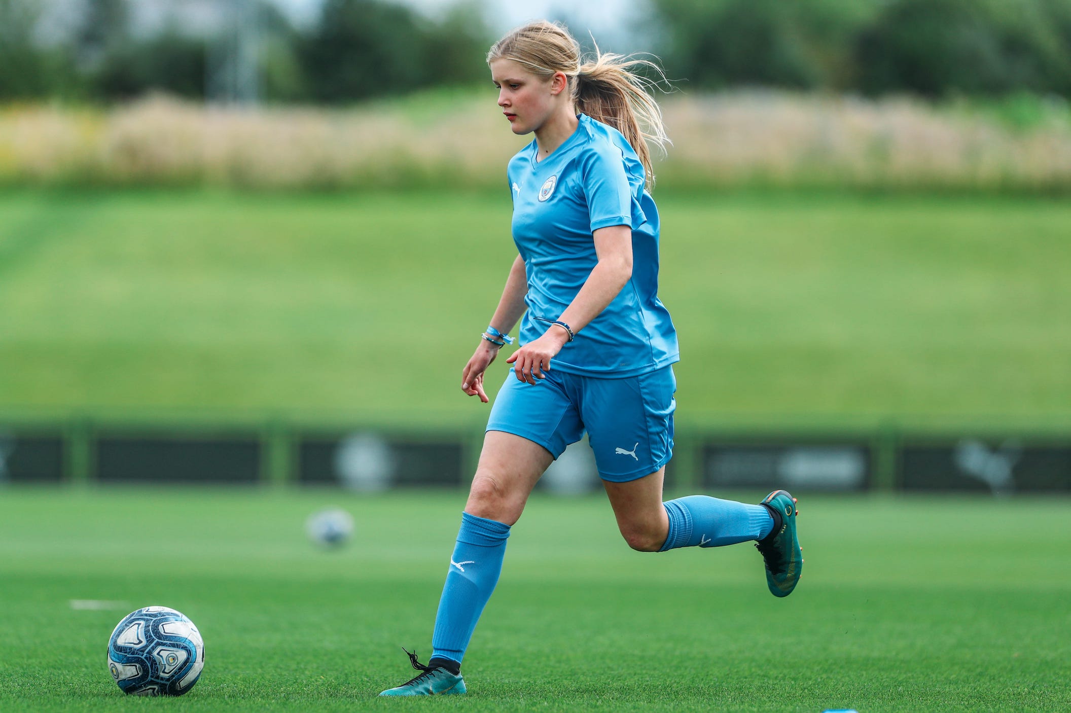 A teenage girl running after a football. She has blonde hair and is wearing a Manchester City Football School kit.