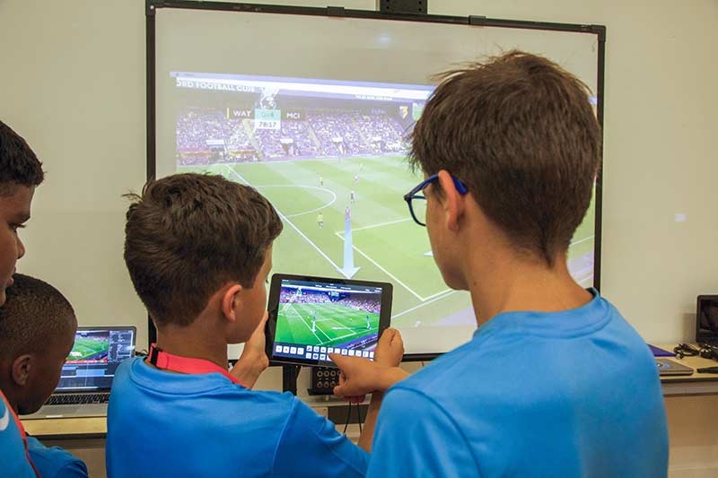 Two boys watching a football match on a tablet. In the background is a large TV screen that also features a football match.