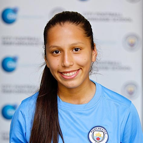 A young girl smiling for a photo. Her ponytail is sat over her shoulder and she is wearing a Manchester City Football School top. The background is blurred. 