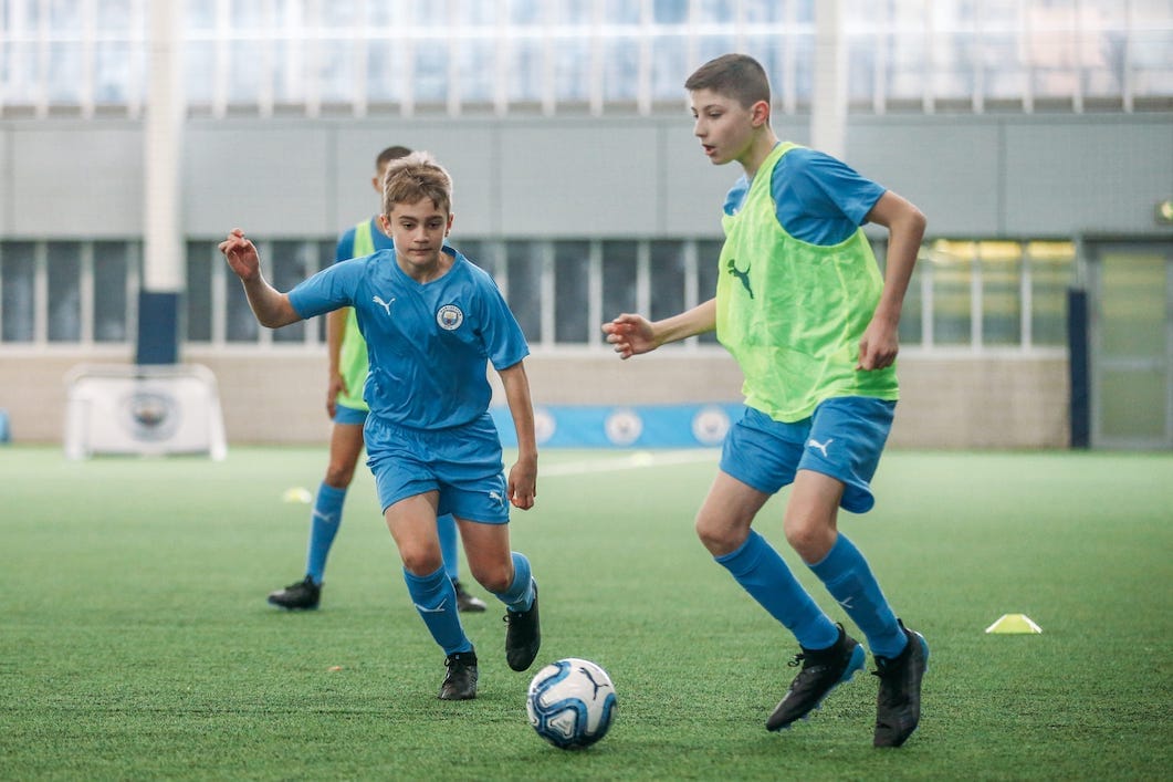 Two boys at City Football Language School chasing a football. Both are wearing a blue football kit. One boy is wearing a green bib.