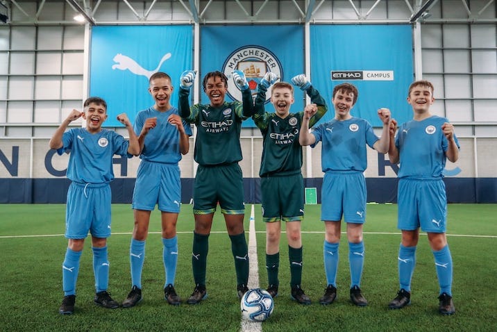 Six young boys at the youth football training camp at Manchester City Football School. They are all lined up in front of a football and cheering, with their hands in the air.