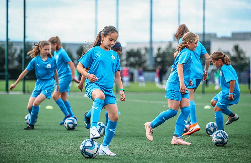 A group of young girls on the Football Development Program at Manchester City Football School. There are seven girls in the photo and each one is focused on dribbling a football.