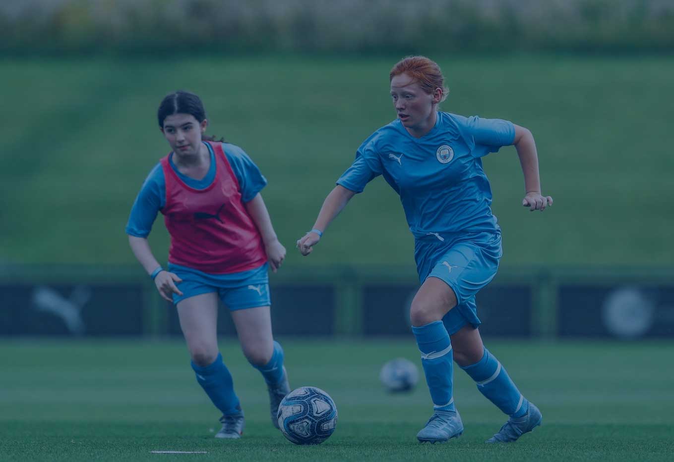 Two young girls chasing a football. Both are wearing a blue Manchester City Football School kit. One girl is wearing a red bib with a puma logo.