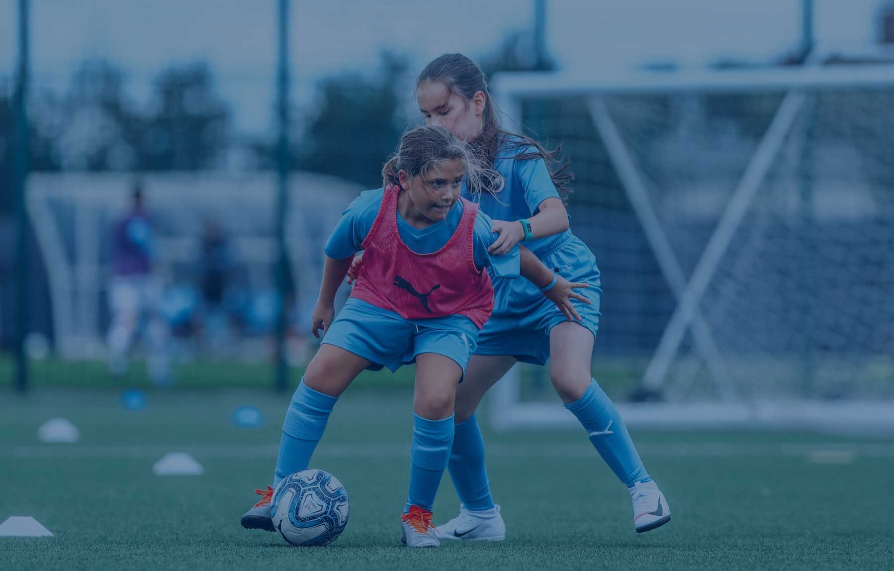 Two young girls in the Football Development Program at Manchester City Football School. The first girl is wearing a red bib and is in possession of the football while the second girl is behind her.