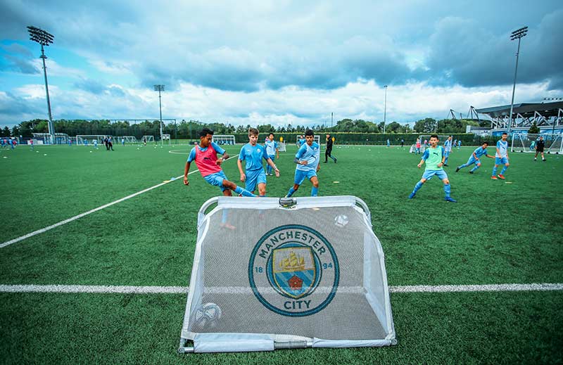 A group of boys running towards a goal at Manchester City Football School. In the foreground of the photo is a goal post with the Man City Football School logo on it