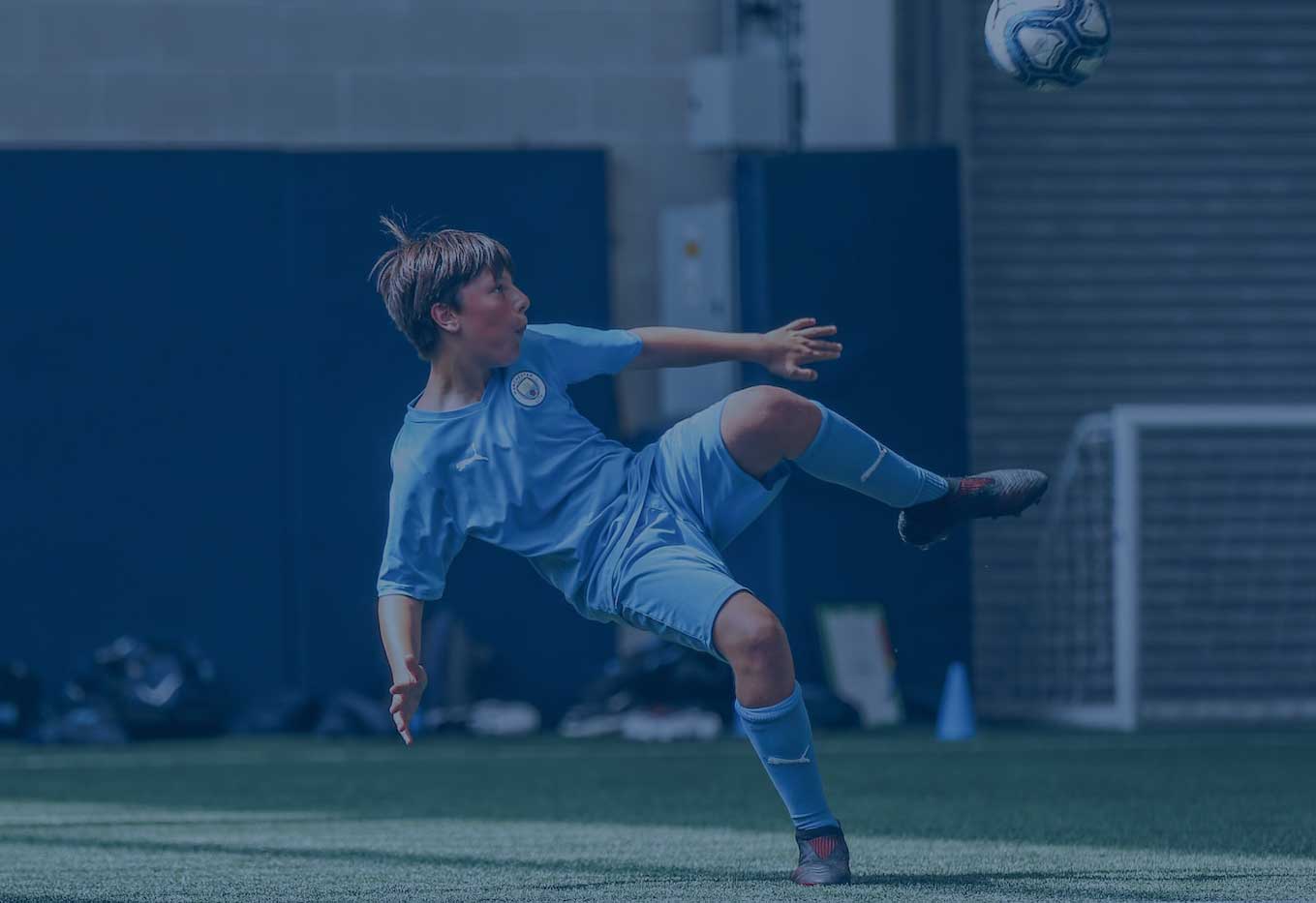 A young boy kicking a football. He has brown hair and is wearing a Manchester City Football School kit.