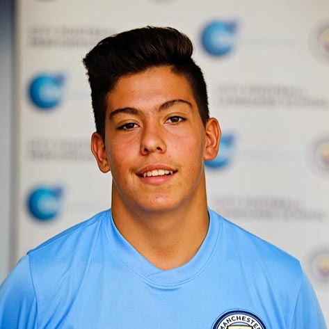 A young boy with dark hair is smiling for a photo. He is wearing a Manchester City Football School top. The background is blurred.