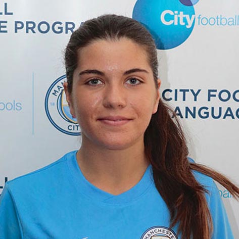 A young girl is posing for a photo. She has her brown hair tied back and is wearing a Manchester City Football School top. In the background is a wall which reads ‘City Football Language School'