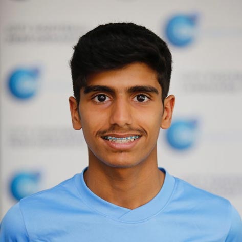 A young boy with dark hair and braces is posing for a photo. He is wearing a Manchester City Football School top. The background is blurred.