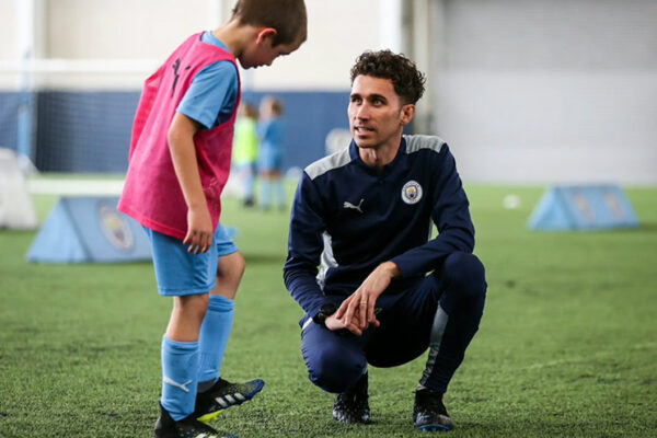 A male football coach kneels to talk to a young football player