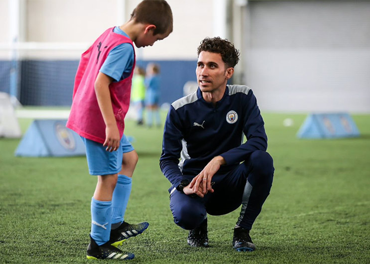 A male football coach kneels to talk to a young football player