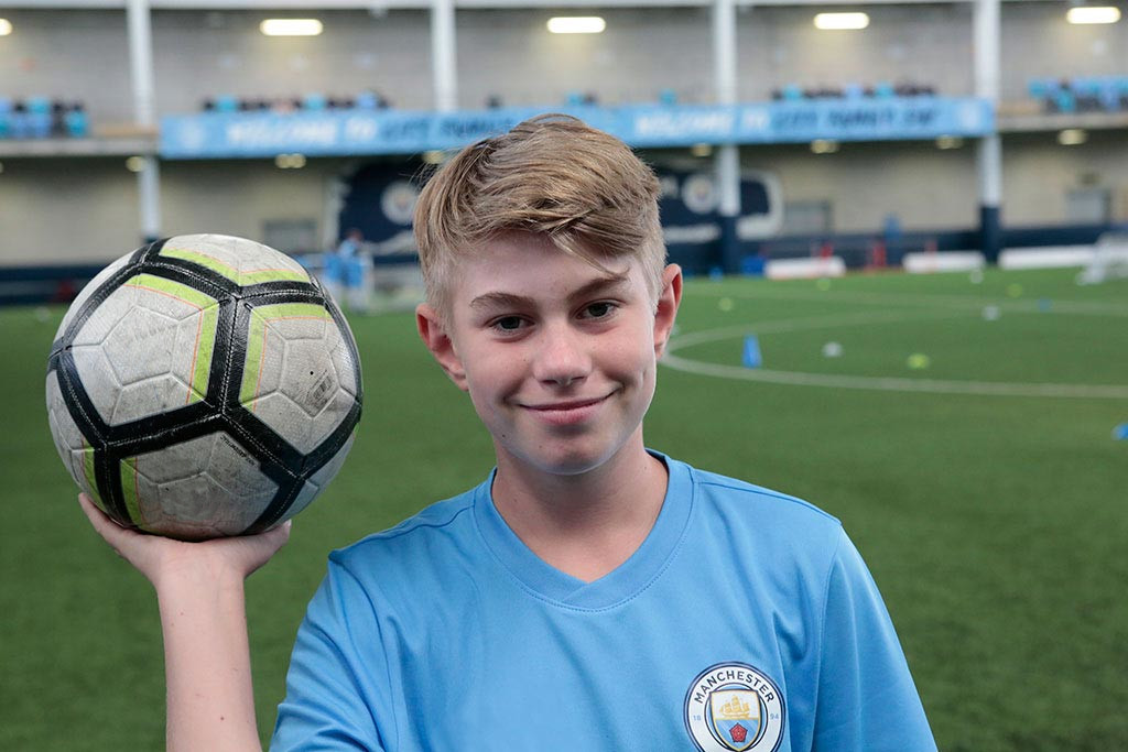 A young boy with blond hair standing on a football pitch and balancing a football in his hand. He is wearing a Manchester City Football School kit.